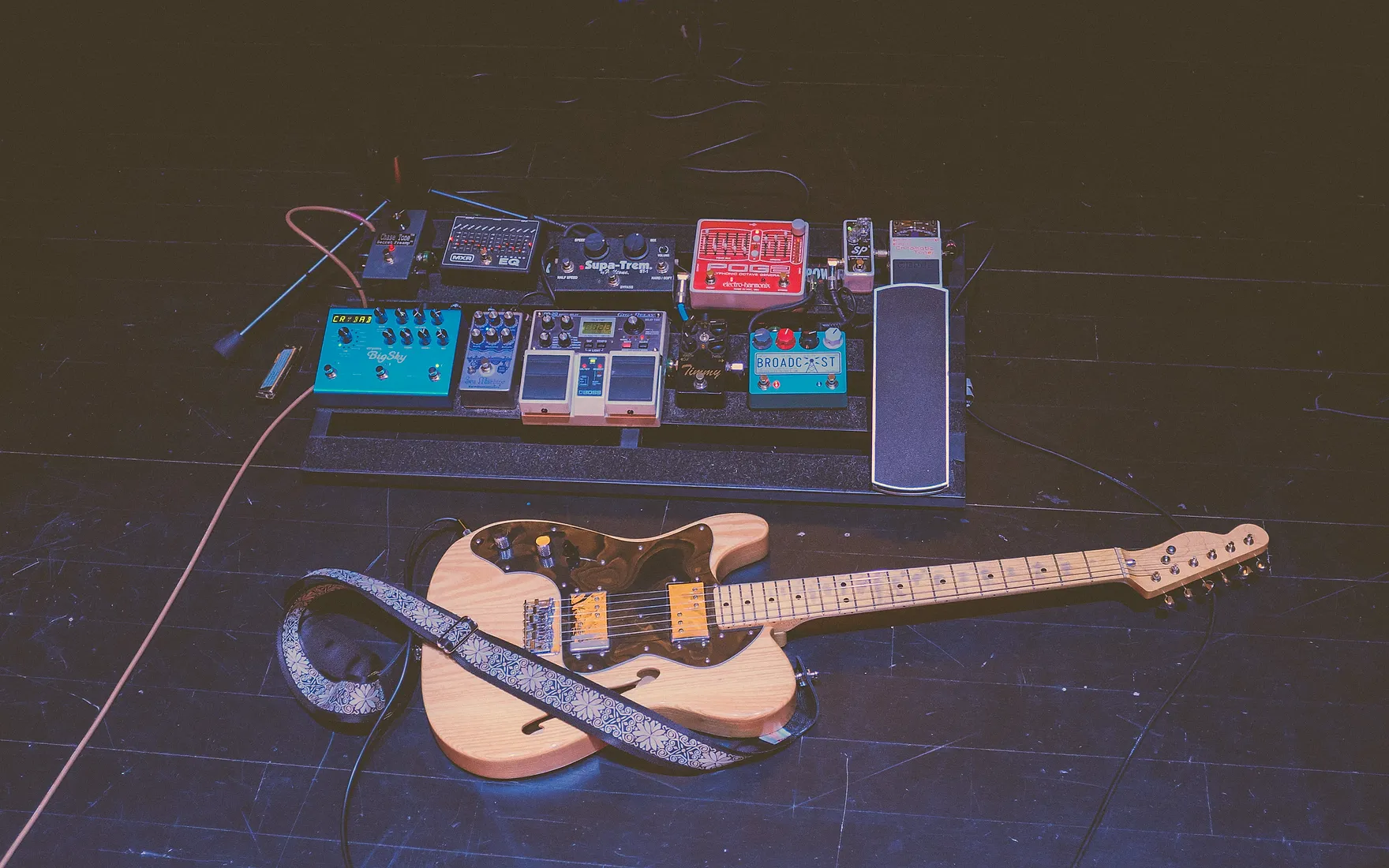 An electric guitar resting on a darkened stage beside a pedalboard of drive, delay, and reverb pedals